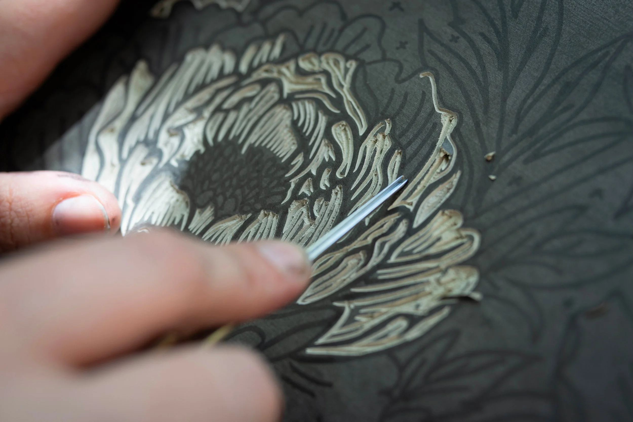 Close-up of a person's hand carving a detailed floral design into a dark linoleum surface.