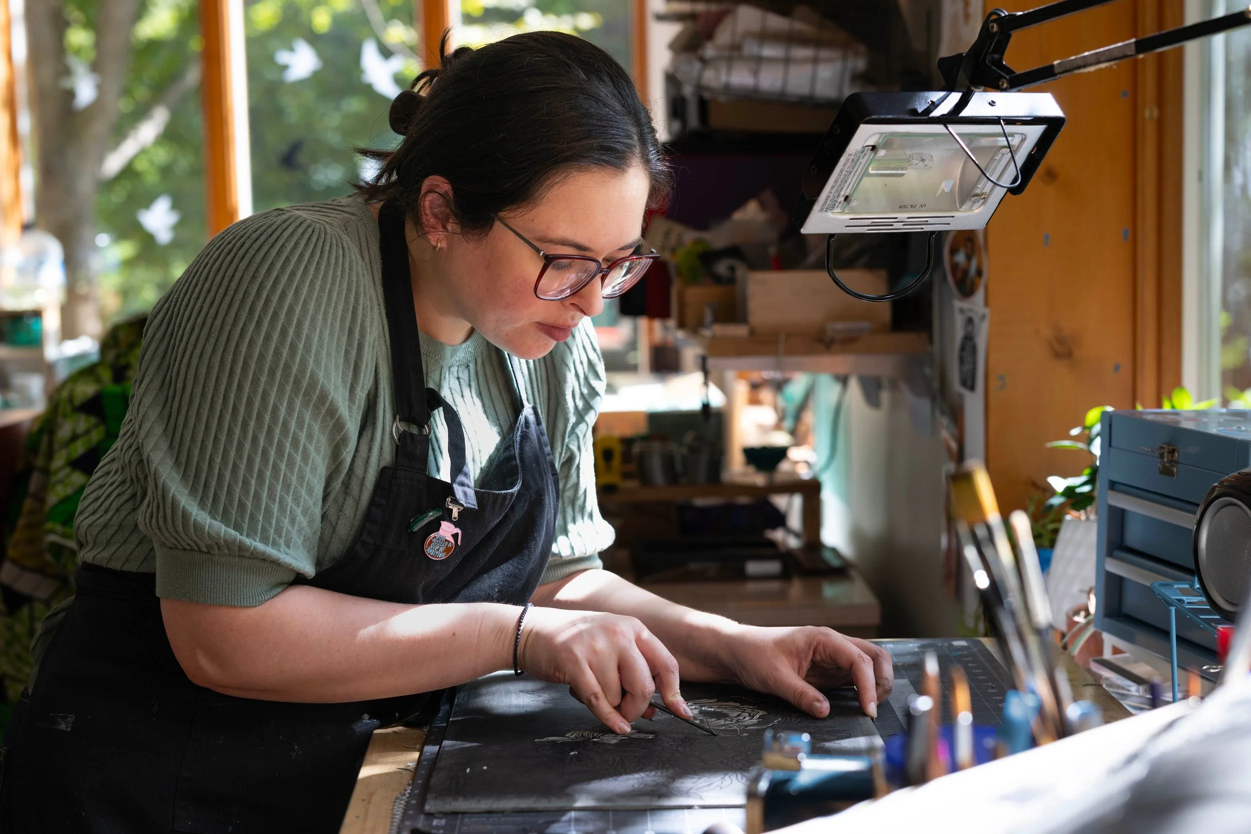 Woman with dark hair, glasses, and a black apron working on a detailed craft project at a workbench in a well-lit workshop with large windows.