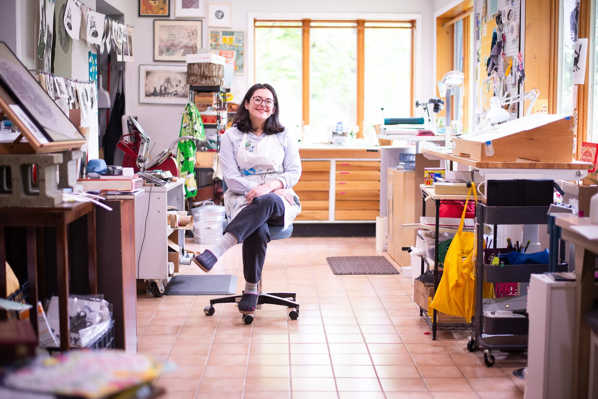 A woman with glasses and black hair smiling while sitting on an office chair in a craft or art studio. The studio is filled with artwork, sketches, and art supplies on shelves and walls, with large windows in the background letting in natural light.