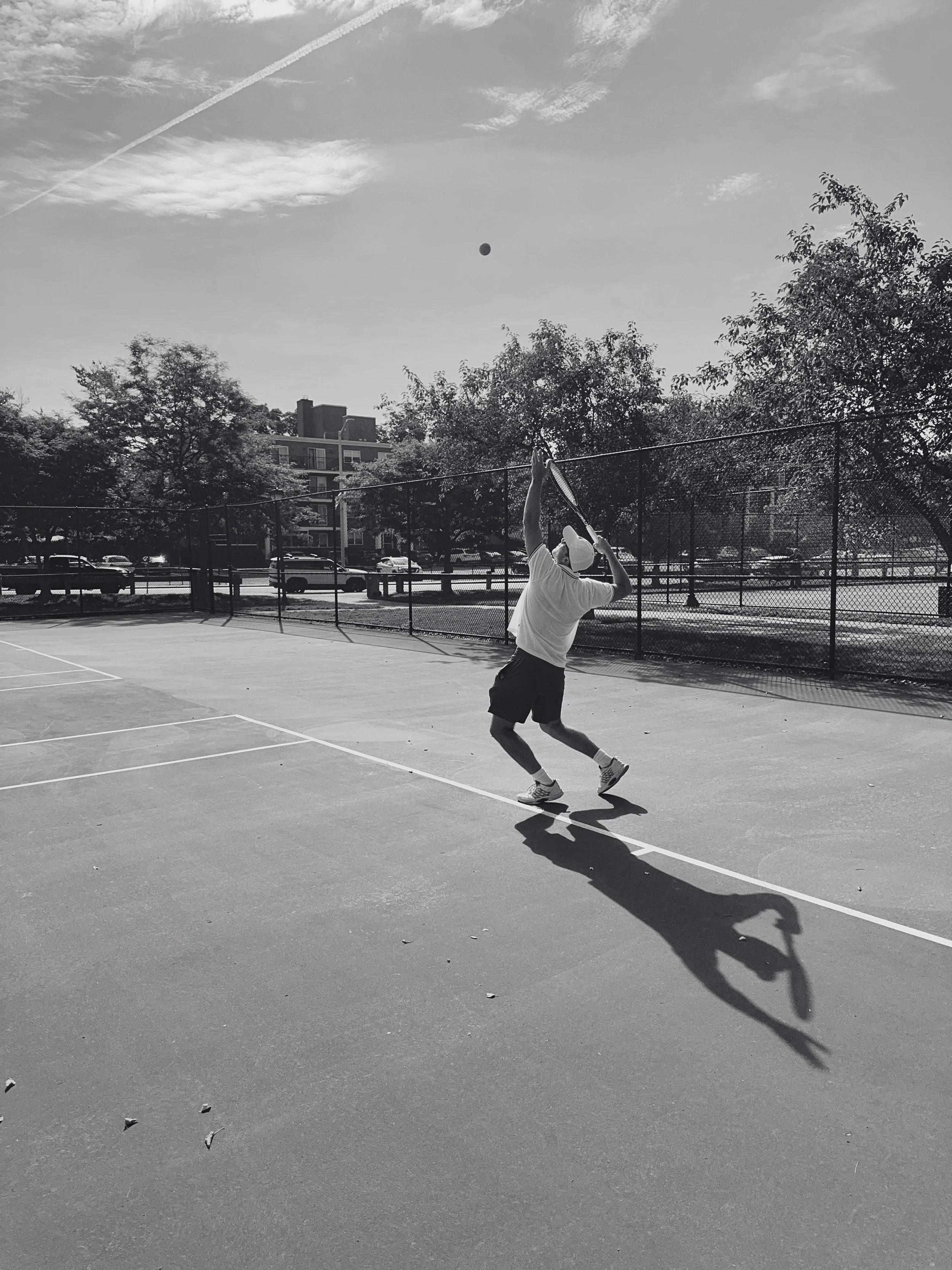 A man playing tennis on an outdoor court with a tennis ball in the air, trees, cars, and buildings in the background, under a partly cloudy sky.