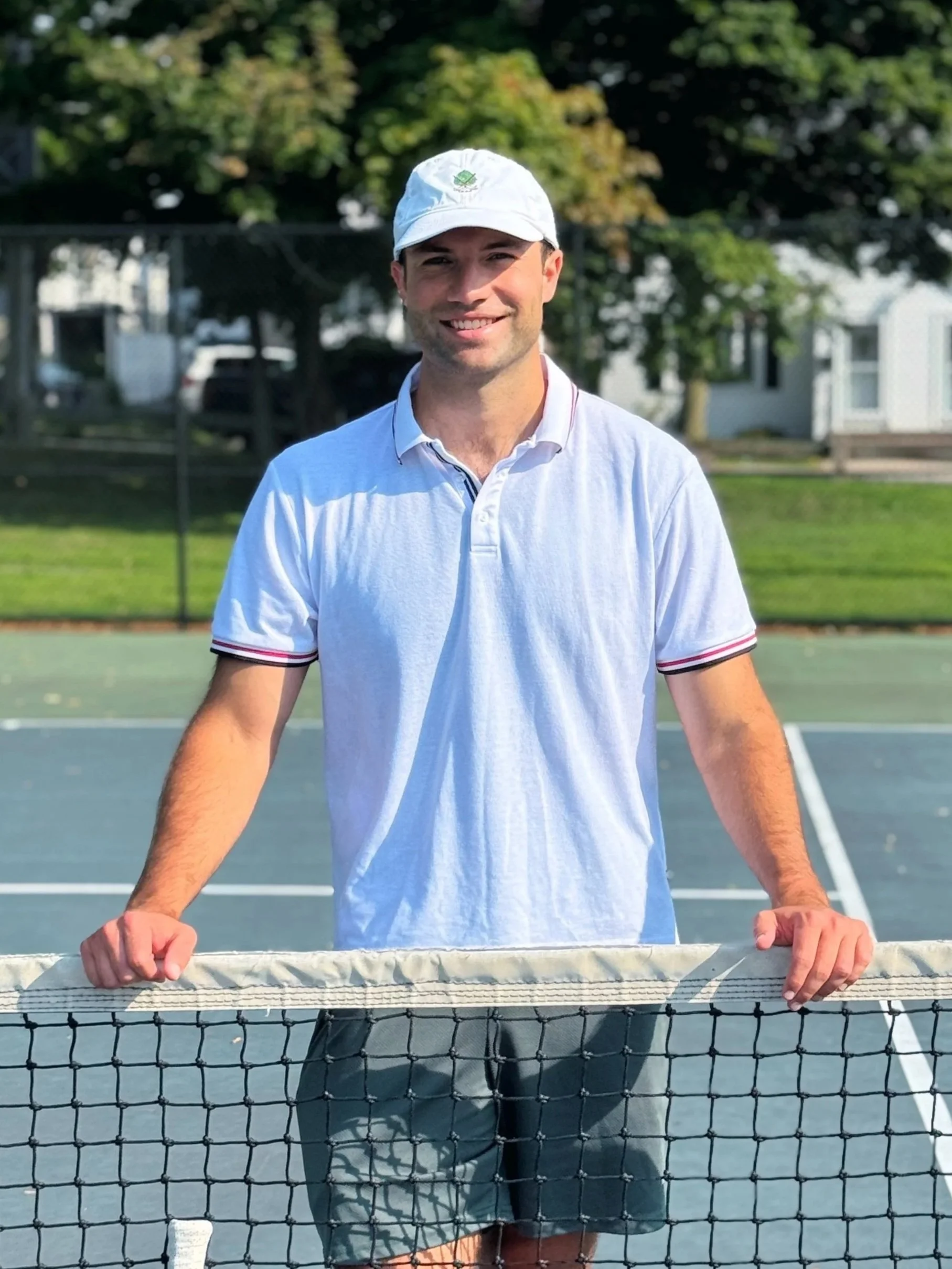 A man standing behind a tennis net on a tennis court, smiling, wearing a white cap and a light-colored polo shirt, with trees and houses in the background.