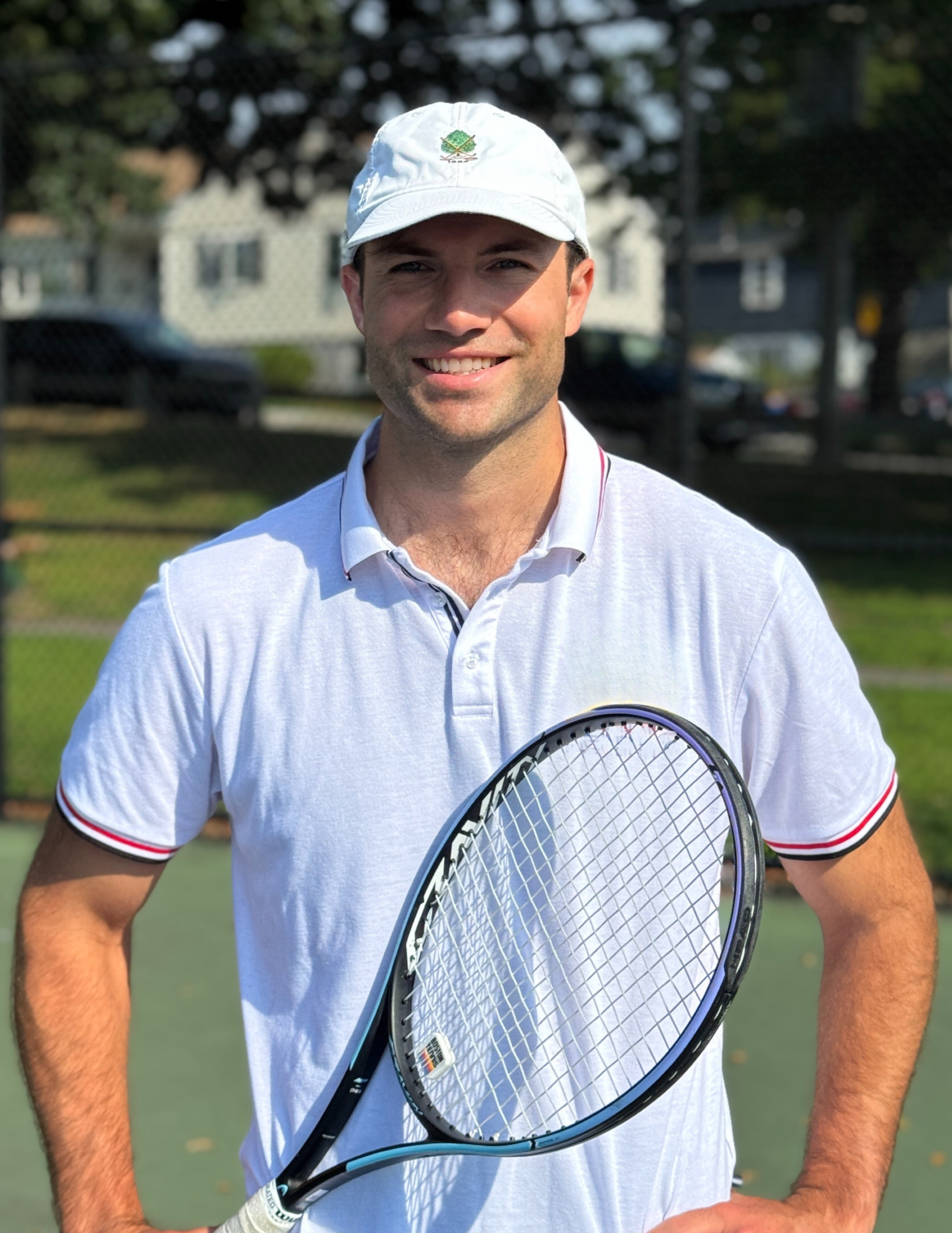A smiling man in a white tennis shirt and white cap, holding a tennis racquet, standing on a tennis court with trees and houses in the background.