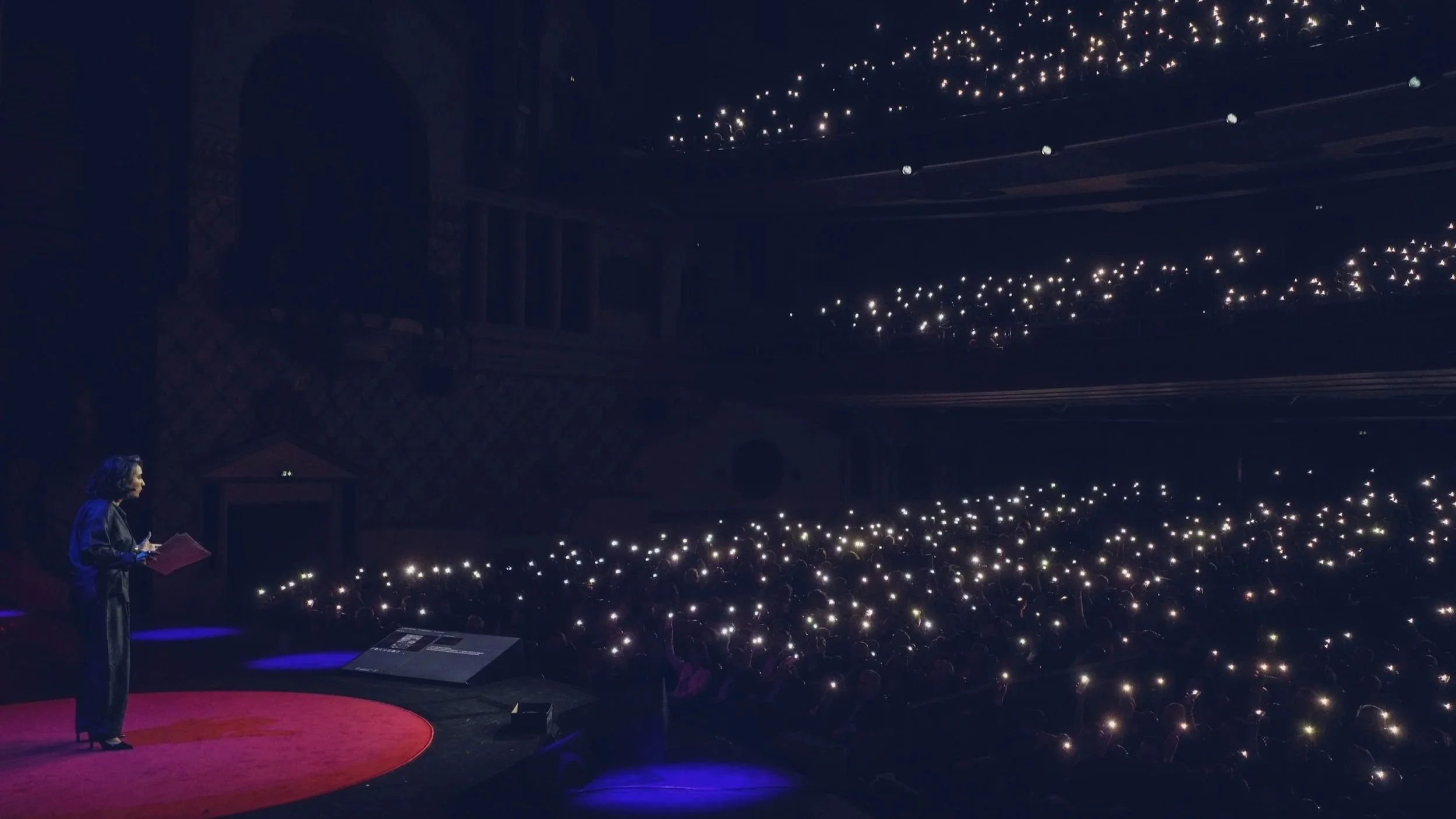 Une femme parle sur scène au TEDx, avec une foule dans une salle sombre utilisant des lumières de téléphone, dans un lieu avec des arches en pierre et un plafond orné de petites lumières fixes.