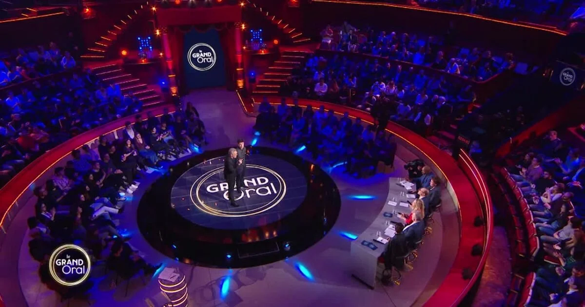 Une scène de spectacle télévisé dans une salle circulaire avec un public assis autour d'une scène centrale où deux personnes parlent, projecteur lumineux et logo 'Le Grand Oral' visible sur le sol et un panneau en arrière-plan.