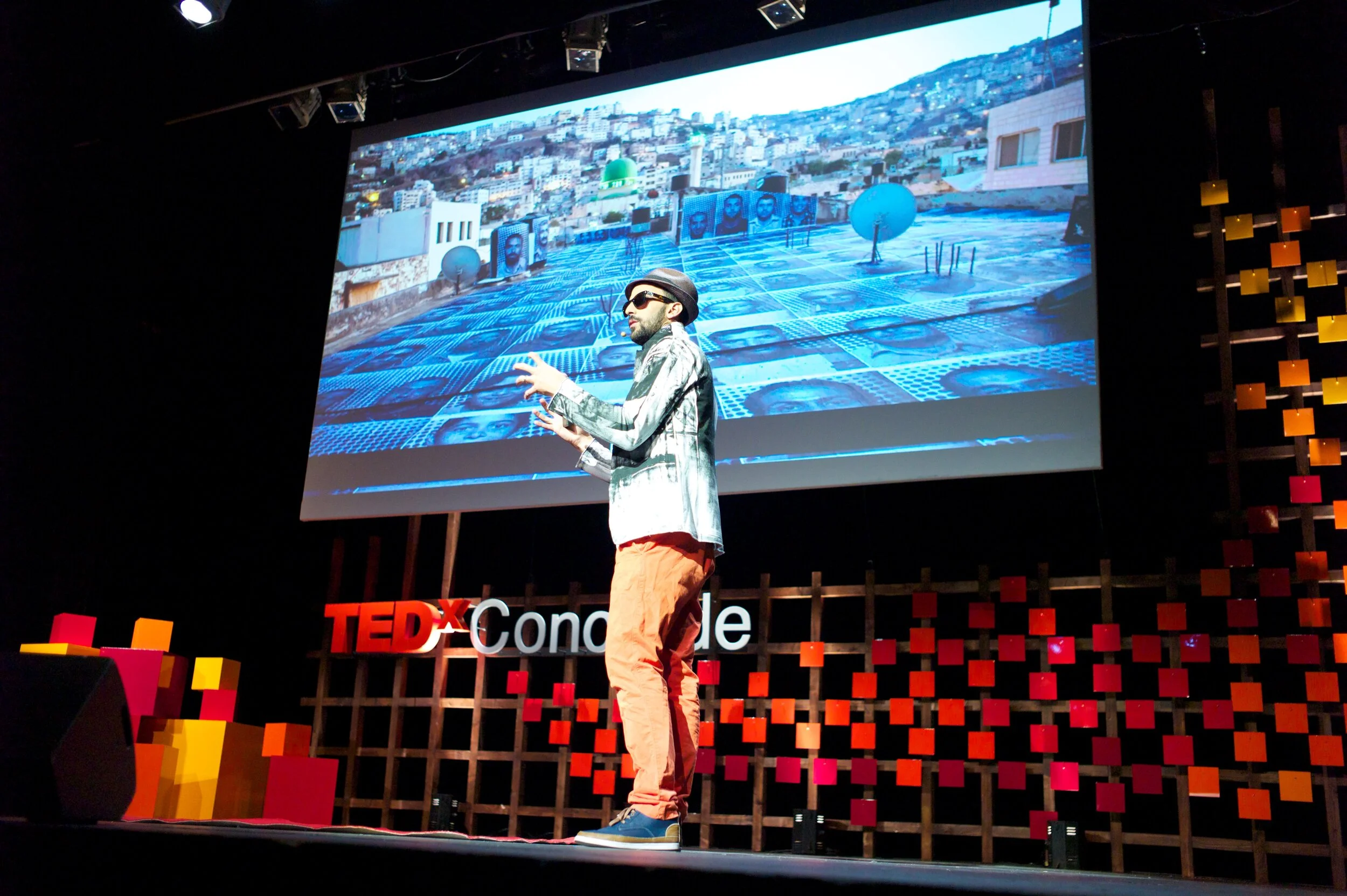 Un homme en vêtements décontractés, portant des lunettes de soleil et un chapeau, parle sur scène lors d'une conférence TEDx, avec un grand écran derrière lui affichant une image d'une ville avec des photographies de personnes sur une surface en toit