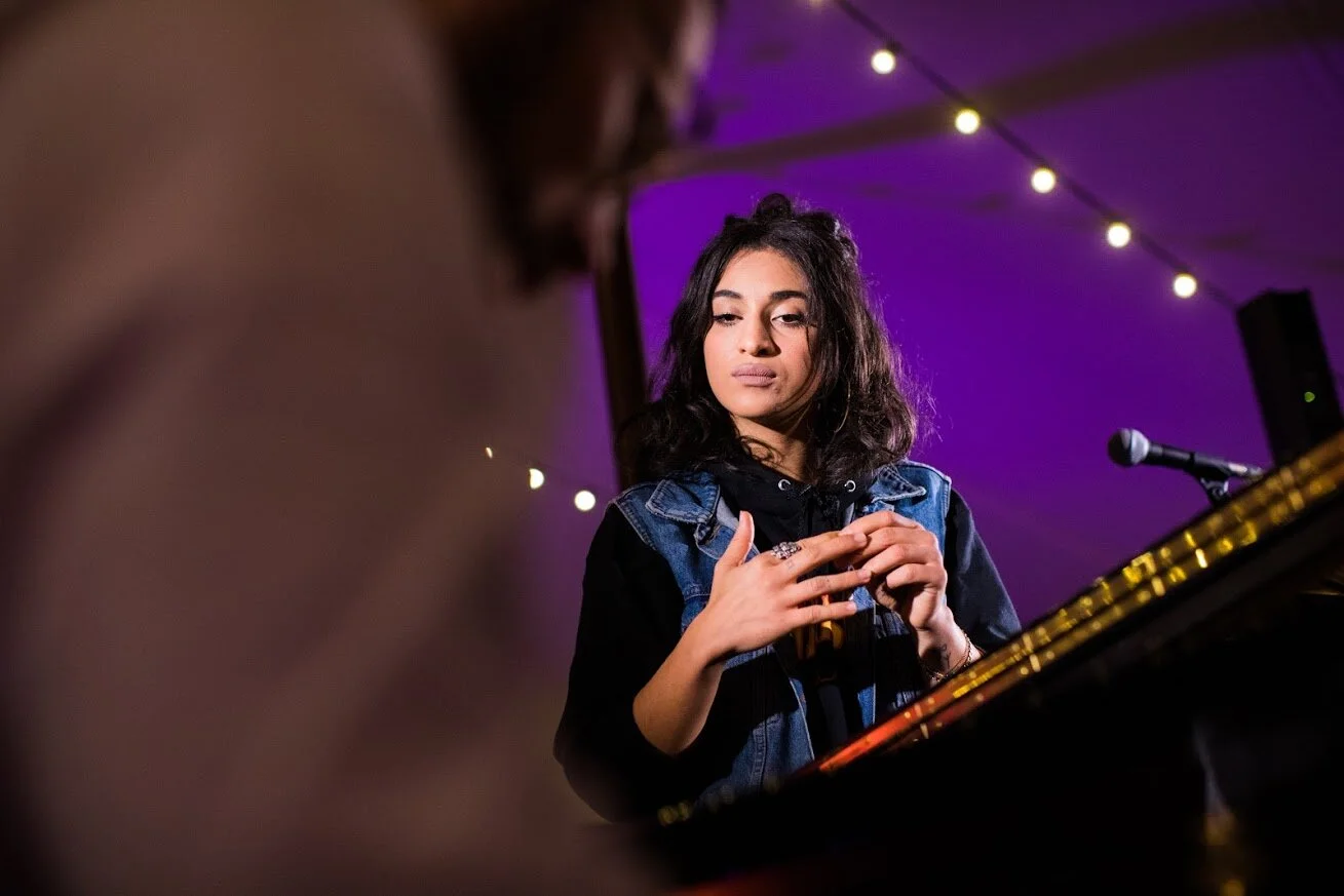 Une jeune femme joue du piano lors d'une performance musicale dans un lieu avec des lumières tamisées violet et blanc.