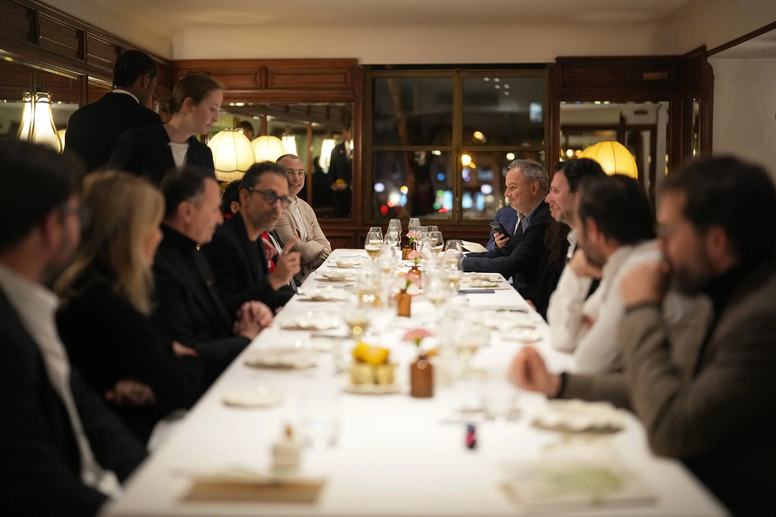 Groupe de personnes assises autour d'une longue table dans un restaurant pendant un dîner, avec des serveurs en arrière-plan.
