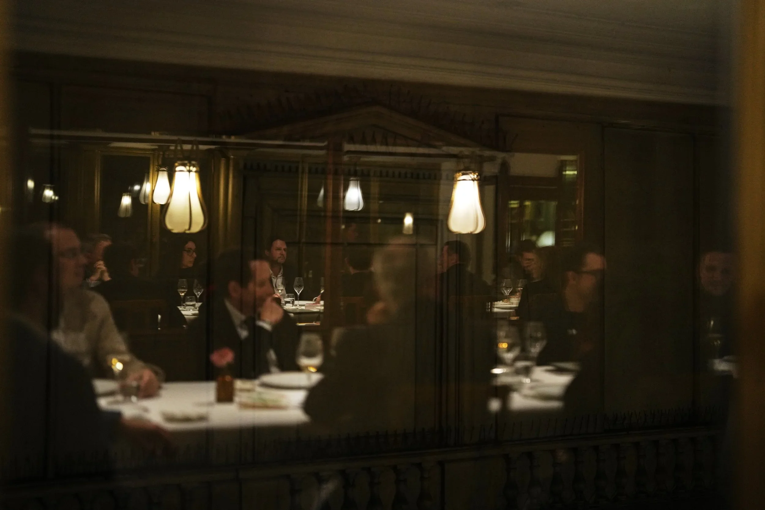 Vue à travers une vitre refletant des personnes assises à une table dans un restaurant ou une salle de réunion, avec des lampes suspendues et un décor élégant.