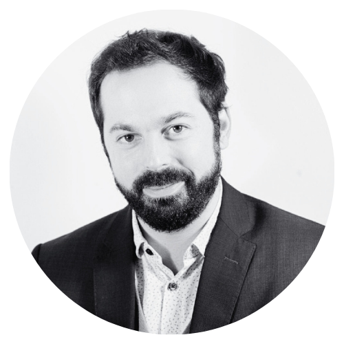 Homme avec barbe et cheveux courts, portant un costume, souriant, fond blanc