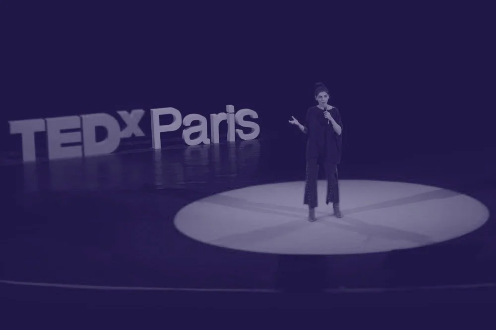 Une personne parlant lors d'un speech au TEDx Paris, sur scène avec un fond sombre et un panneau TEDx Paris à côté.