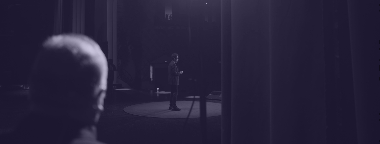 Intervenant seul sur une scène circulaire éclairée, vu depuis les coulisses, donnant une keynote devant un public lors d’une conférence.