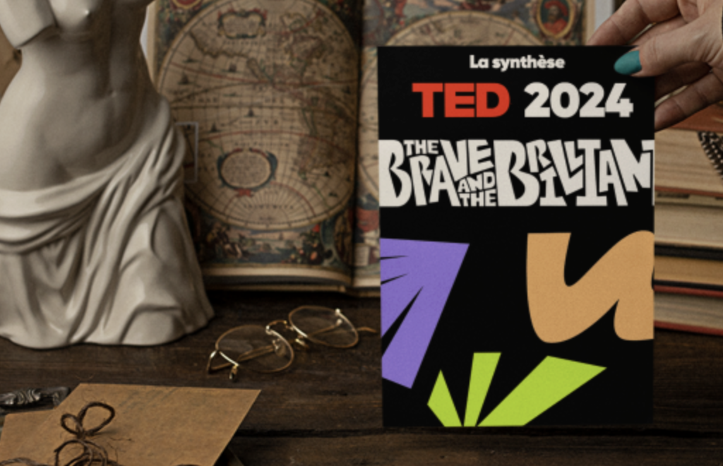 Une brochure TED 2024 intitulée 'The Brave and the Biltan' tenue par une personne, avec des livres anciens, une statue en forme de bust et une paire de lunettes sur un bureau en bois.