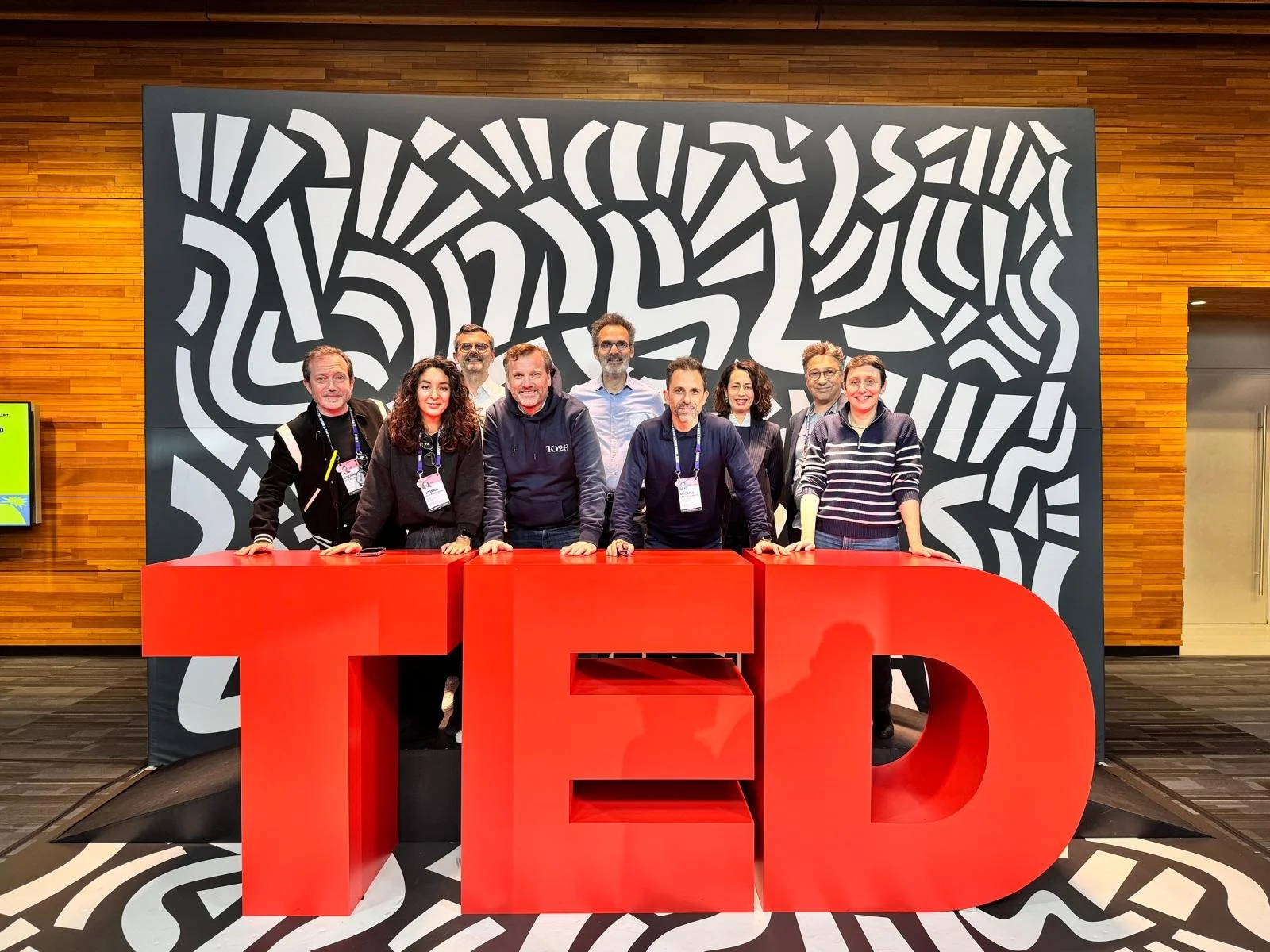 Groupe de personnes posant derrière une grande inscription rouge 'TED' lors d'un événement, avec un mur artistique en noir et blanc en arrière-plan.