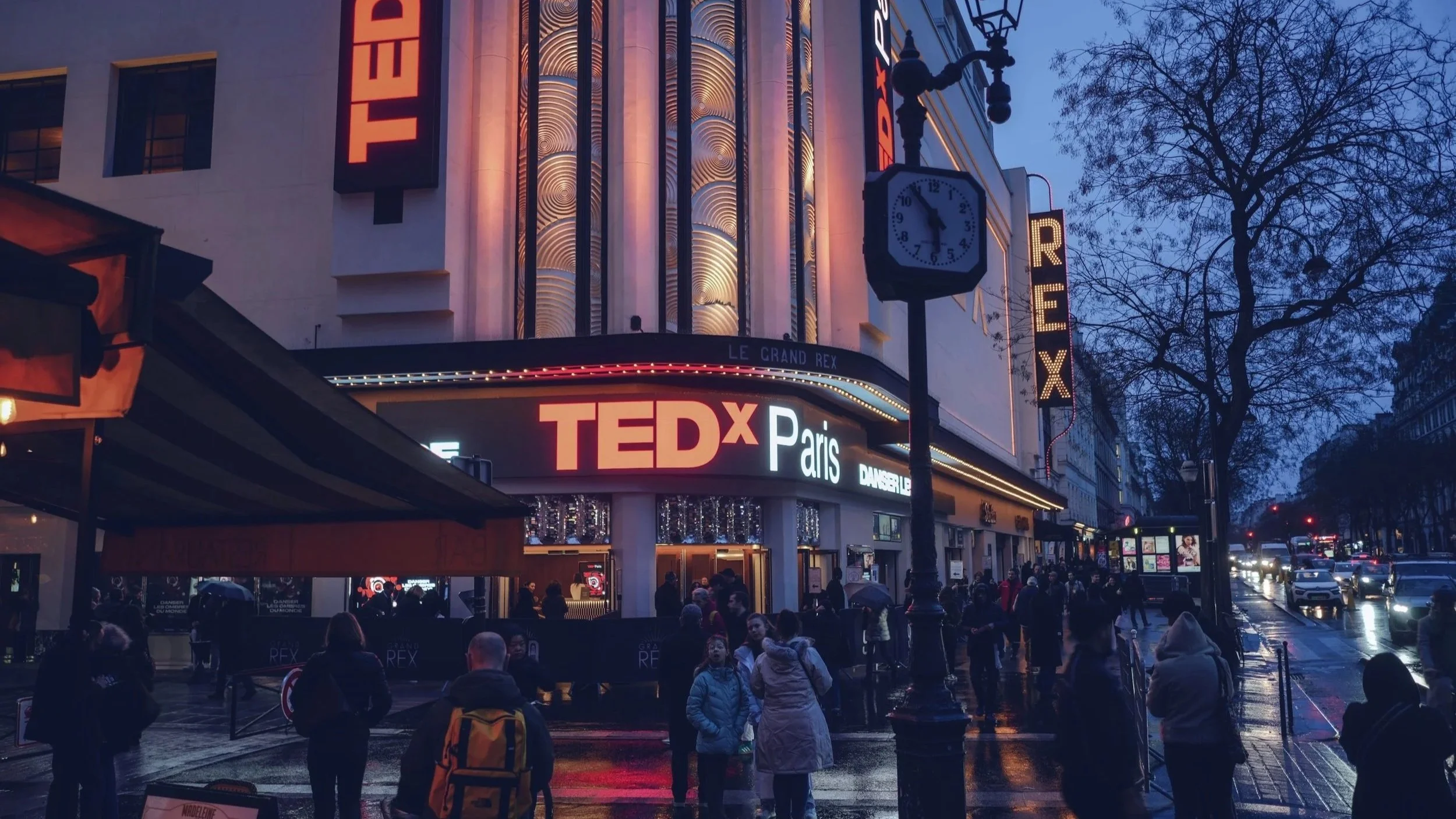 Affiche d'un événement TEDxParis au Grand Rex à Paris, avec une façade illuminée et un clocher d'horloge, peu après le coucher du soleil, dans une rue animée