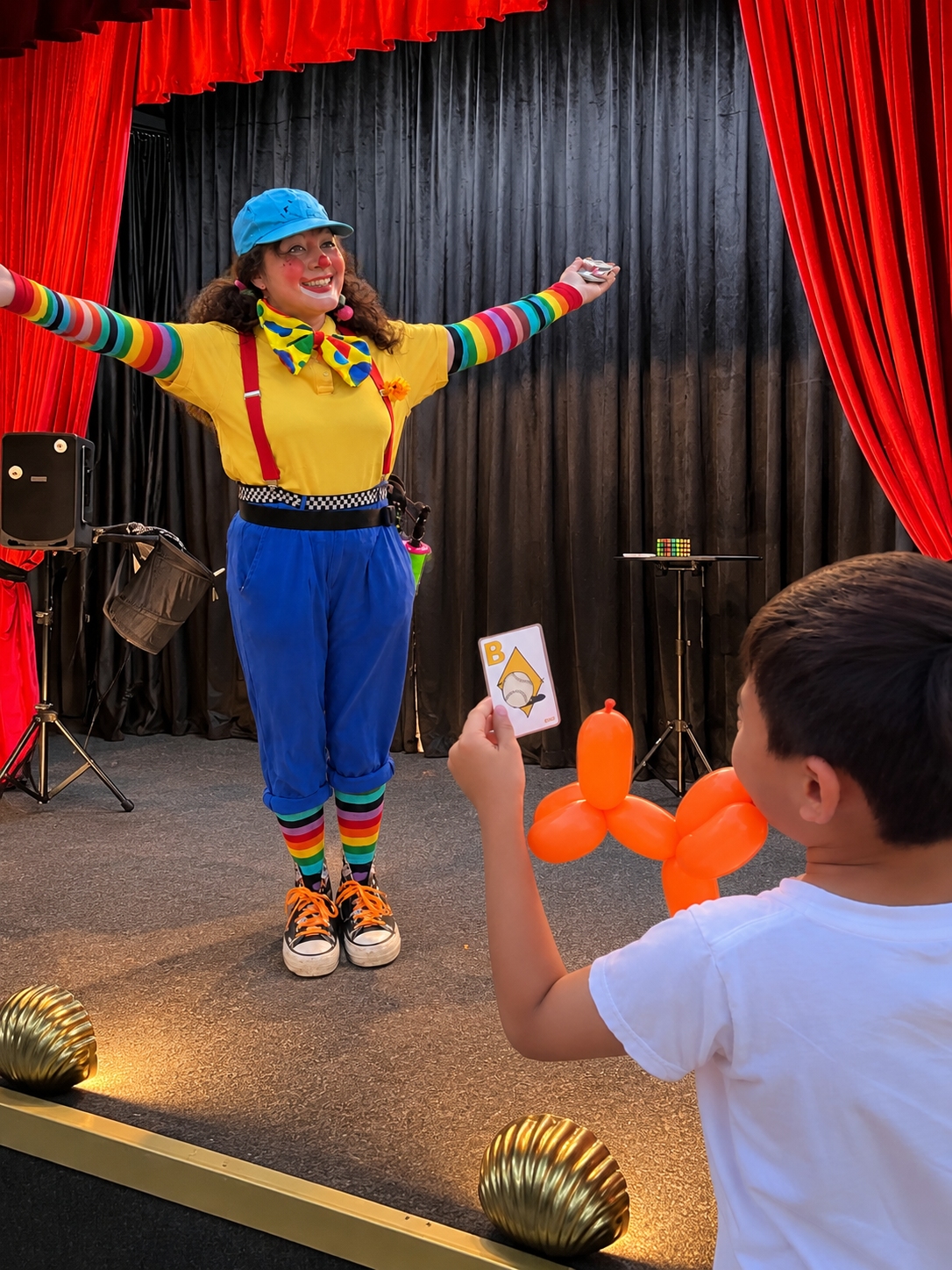 A colorful clown proudly reveals her card prediction on stage during a magic show. The young boy who is her audience participant is holding the card and an animal balloon, and is laughing in delight at the clown's trick.