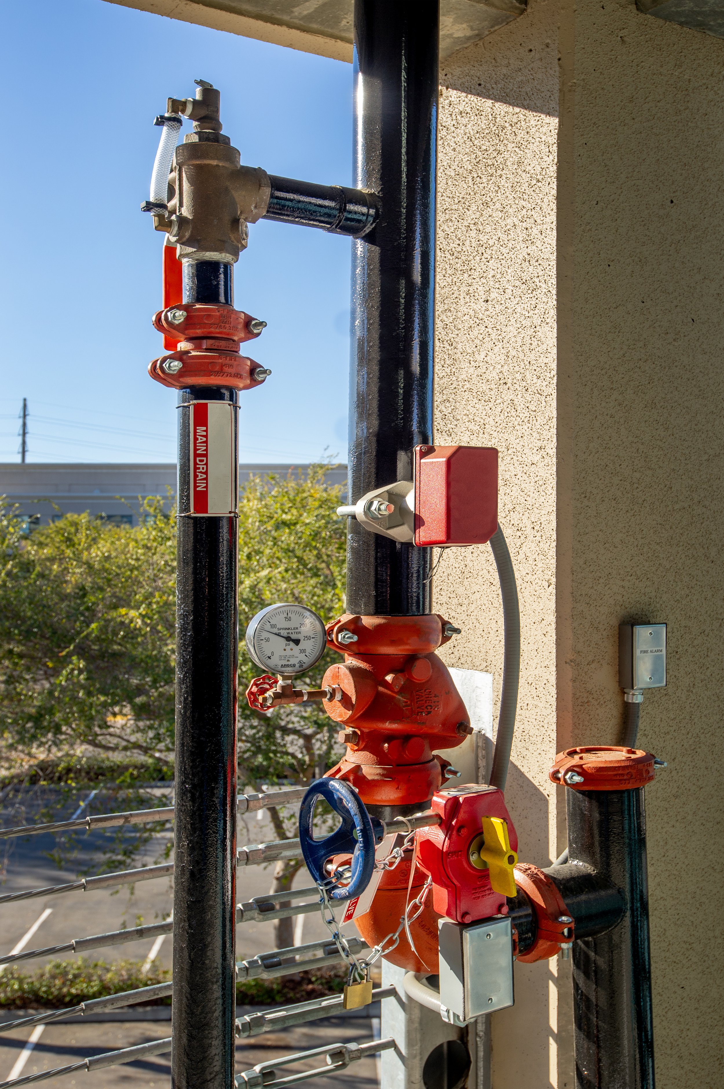 Fire sprinkler system piping with valves, pressure gauge, and alarm box mounted on an exterior wall.