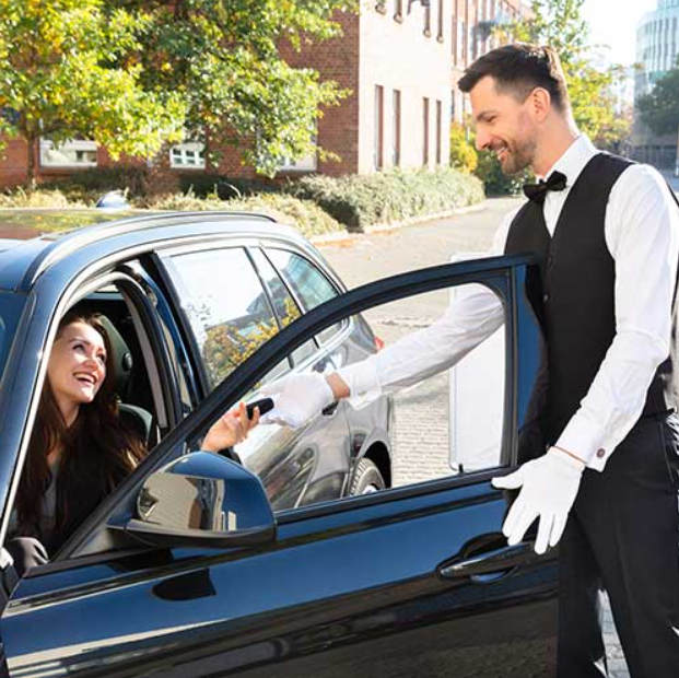 A man in a tuxedo and white gloves handing a key to a smiling woman in the car window on a sunny street.