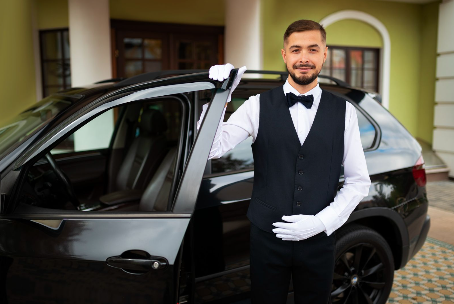 A doorman dressed in a tuxedo with a bow tie and white gloves standing next to a black car, holding the edge of the car's roof.