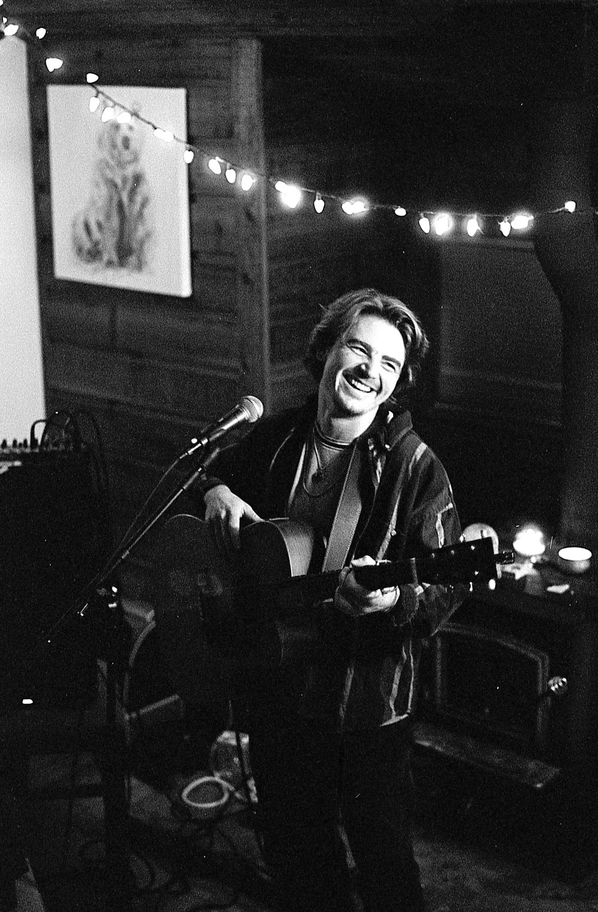 A person with shoulder-length hair is playing an acoustic guitar and singing into a microphone, smiling happily. They are in a cozy indoor setting with string lights hanging above and a painting of a dog on the wall.