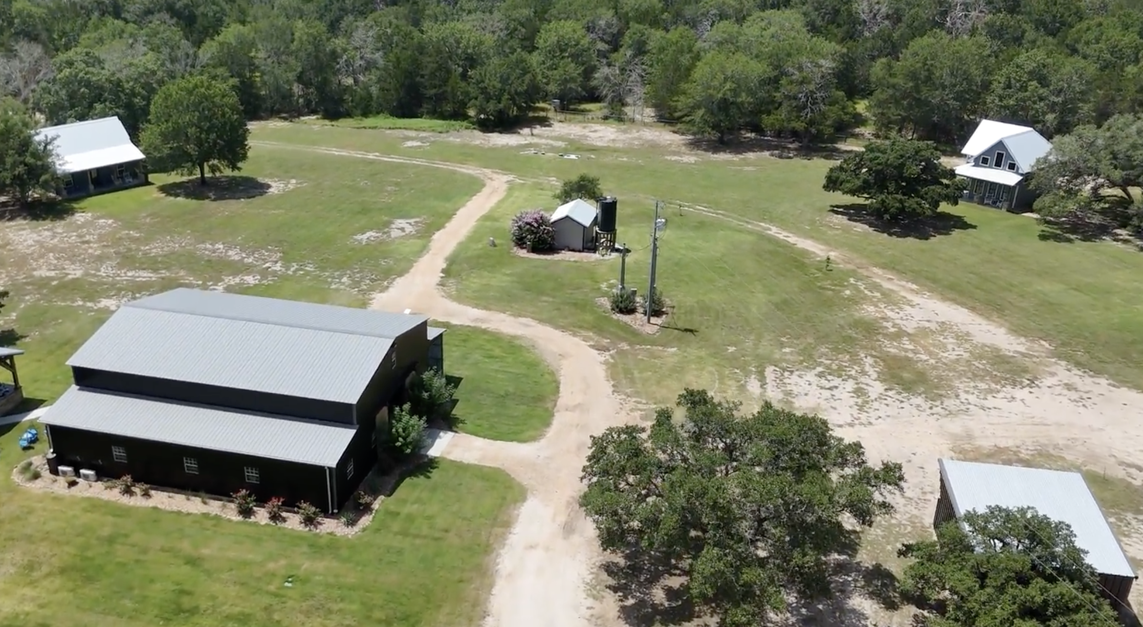 An aerial view of a rural property with several buildings, dirt roads, grassy areas, and trees surrounding the property on a sunny day.