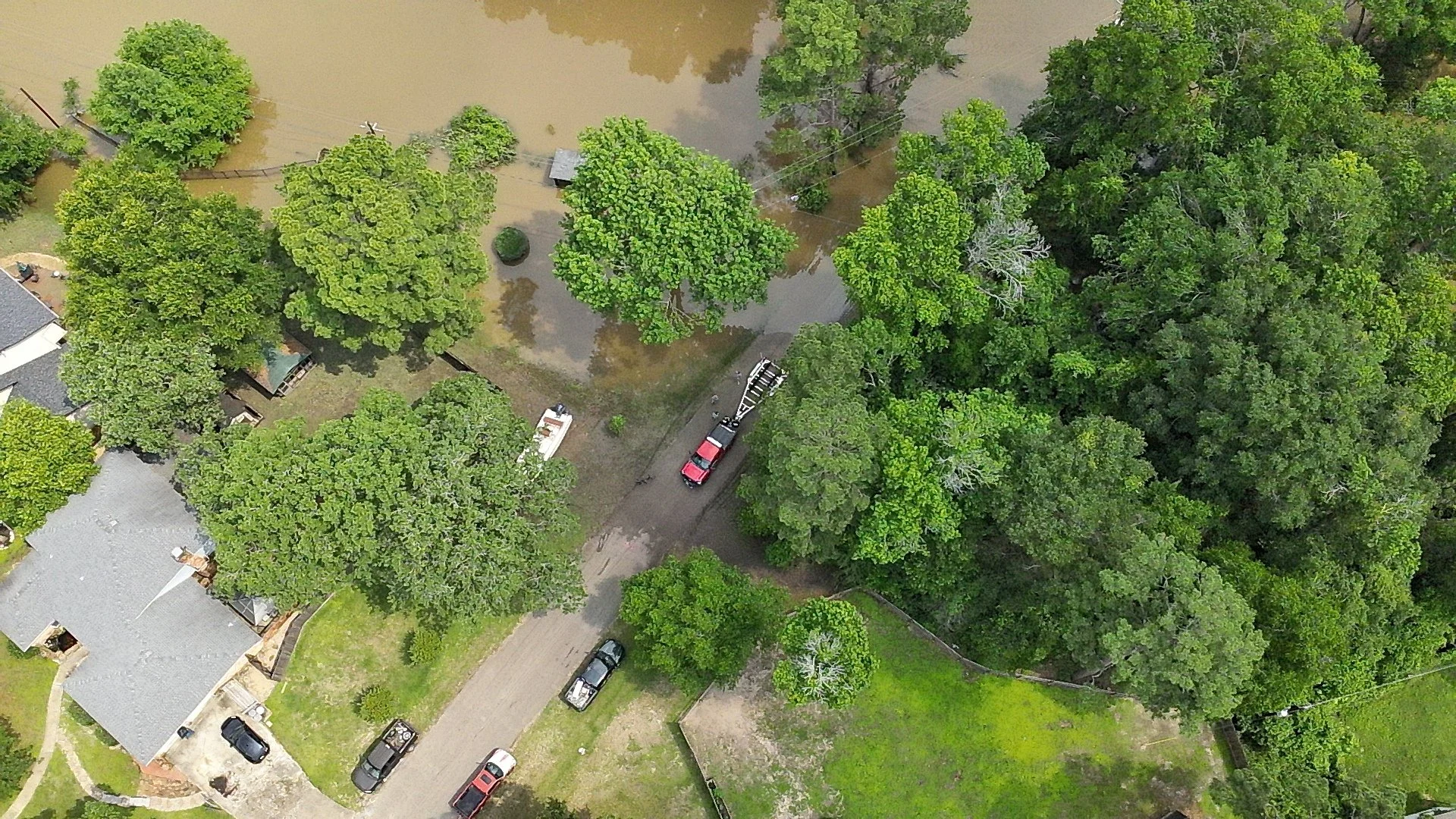An aerial view of a neighborhood affected by flooding, showing water covering yards, roads, and trees, with some parked cars visible.