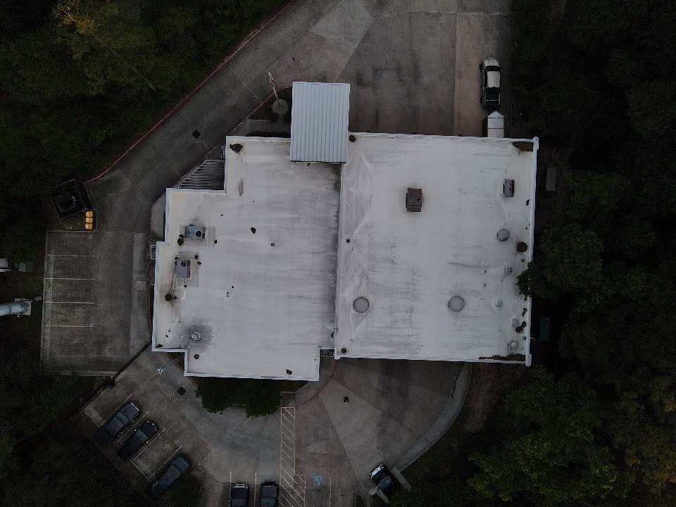 Top-down drone view of a large white building with a flat roof, surrounded by parking lots and trees.