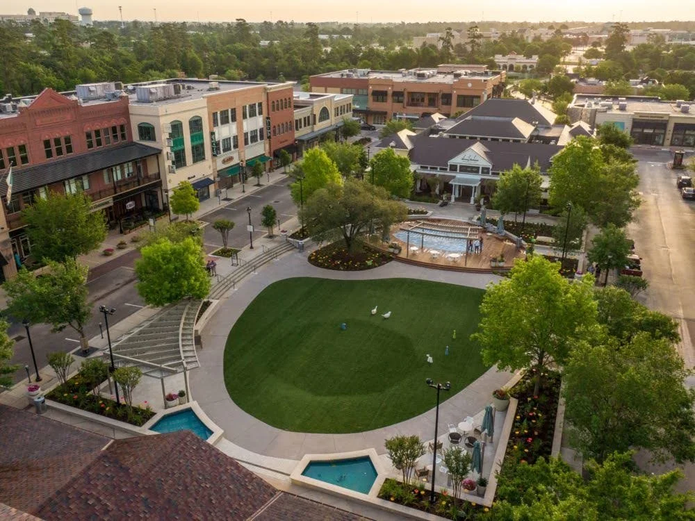 An aerial view of a small square with a grassy area, trees, a fountain, a small pool, and a stage with a pergola, surrounded by shops and buildings in a shopping district.