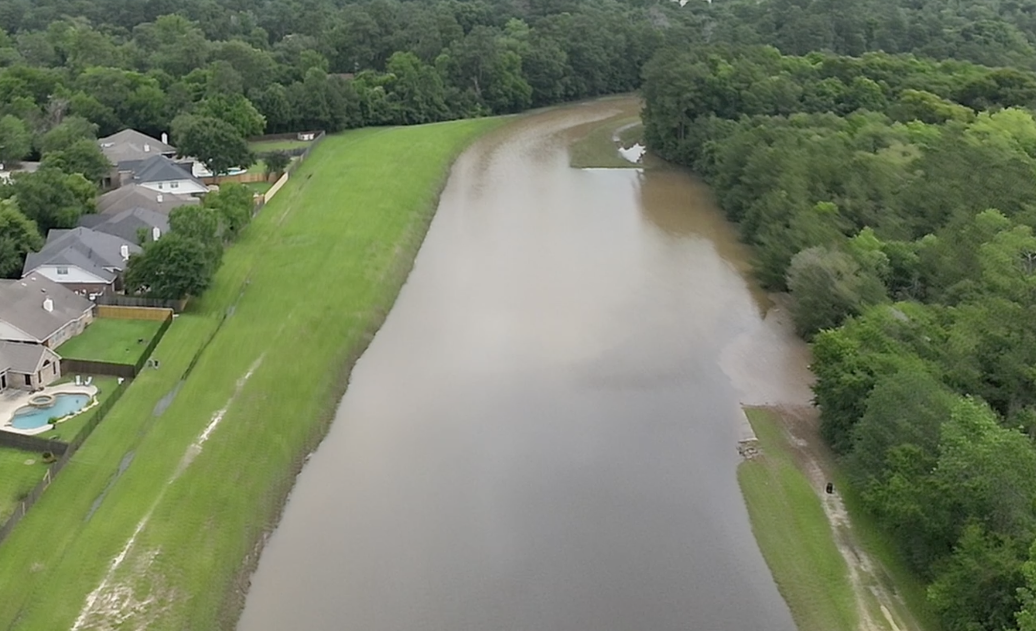 An aerial view of a residential area next to a river with a grassy dam or levee separating the two. The houses have gray roofs, and there is a swimming pool visible in one backyard. Forest surrounds the area.