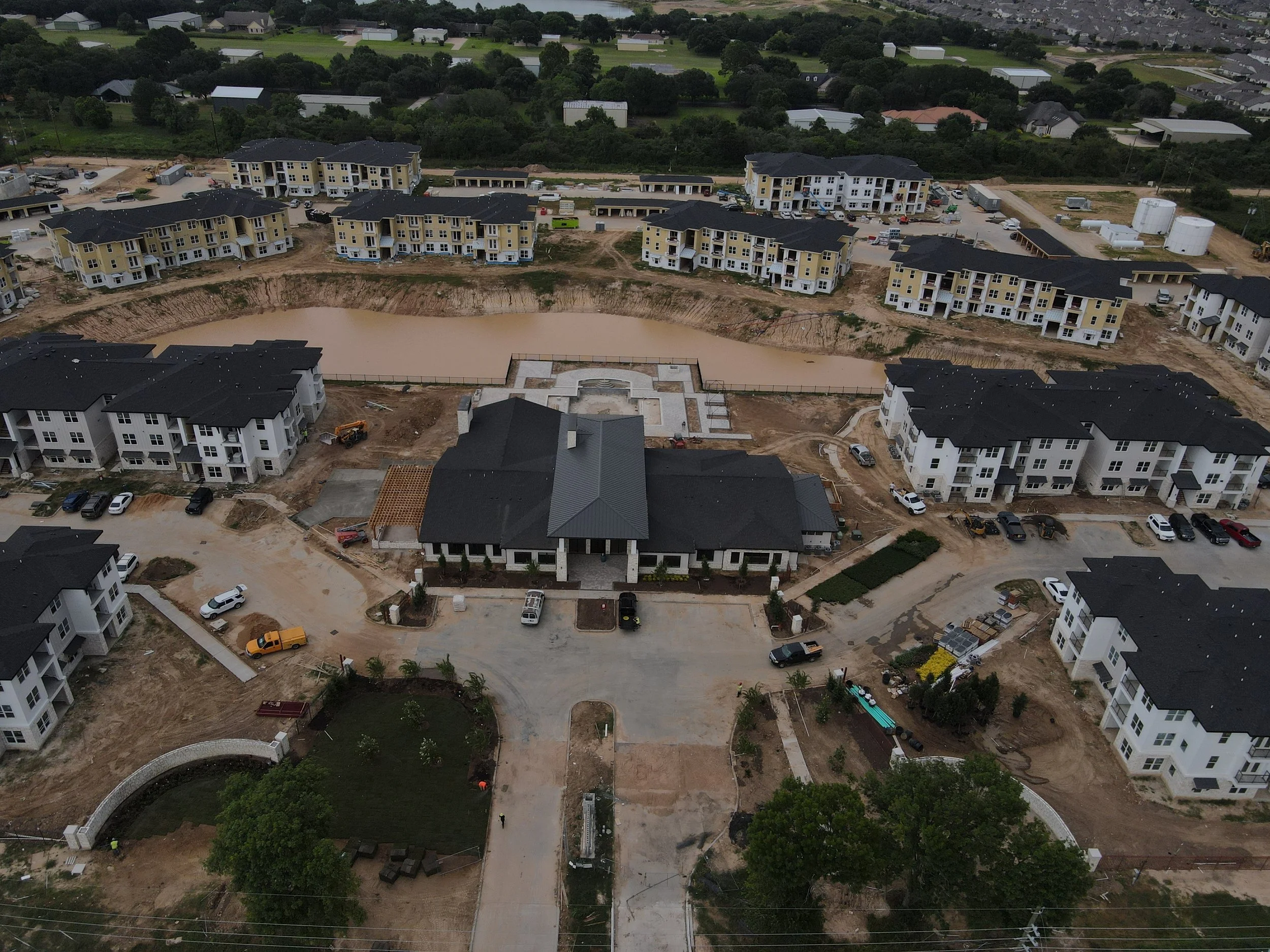 Aerial view of a residential construction site with multiple multi-story buildings in various stages of completion, a pond at the center, construction vehicles, and surrounding greenery.
