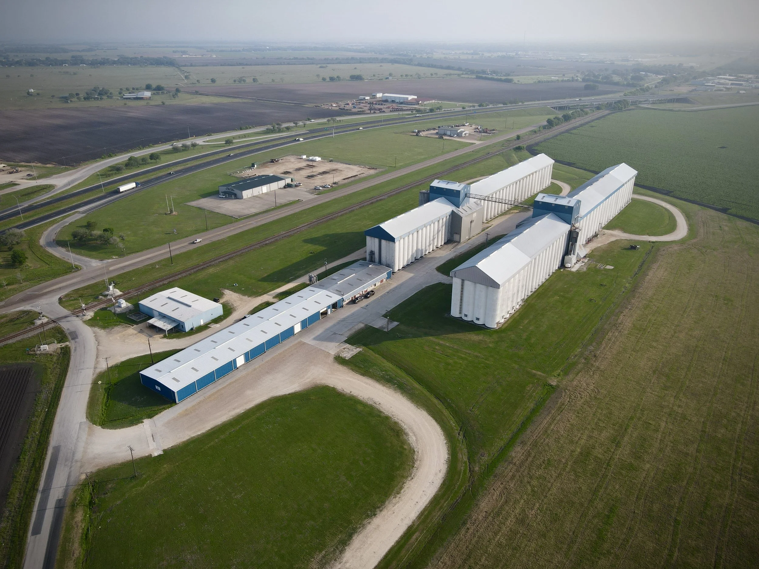 An aerial view of an agricultural facility with large white silos, a blue-roofed building, a parking lot, a highway with cars, and surrounding fields and farmland.