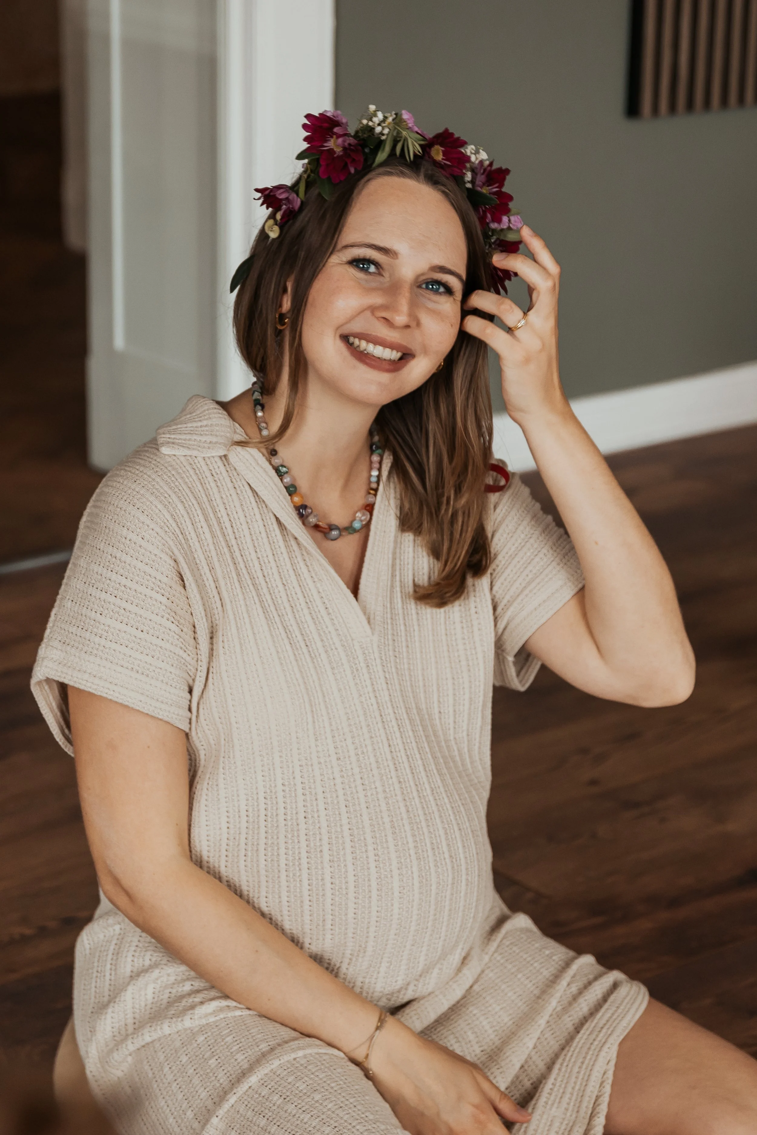 Eine lächelnde junge Frau mit braunen Haaren, die einen Blumenkranz trägt, sitzt auf einem Holzstuhl und hat die Hand am Kopf. Sie trägt eine beige, kurze Ärmel Bluse und eine farbenfrohe Halskette.