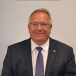 A middle-aged man in a dark suit and tie, smiling, standing against a light-colored background.