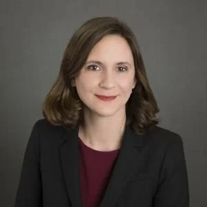 Portrait of a woman with shoulder-length brown hair, wearing a black blazer over a maroon top, smiling in front of a gray background.