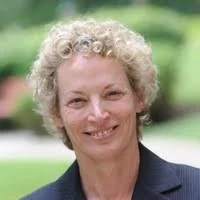 A woman with curly blonde hair smiling outdoors, wearing a dark blazer.