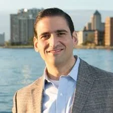 A man in a gray blazer and light blue shirt smiling outdoors with a cityscape and water in the background.