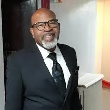 A smiling man wearing glasses, a black suit, white shirt, and a tie, standing indoors near a light-colored wall.