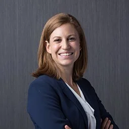 Smiling woman with shoulder-length red hair, wearing a navy blazer and white shirt, standing with arms crossed against a gray background.
