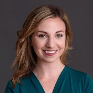 Portrait of a woman with wavy blonde hair wearing a teal top, smiling against a dark background.