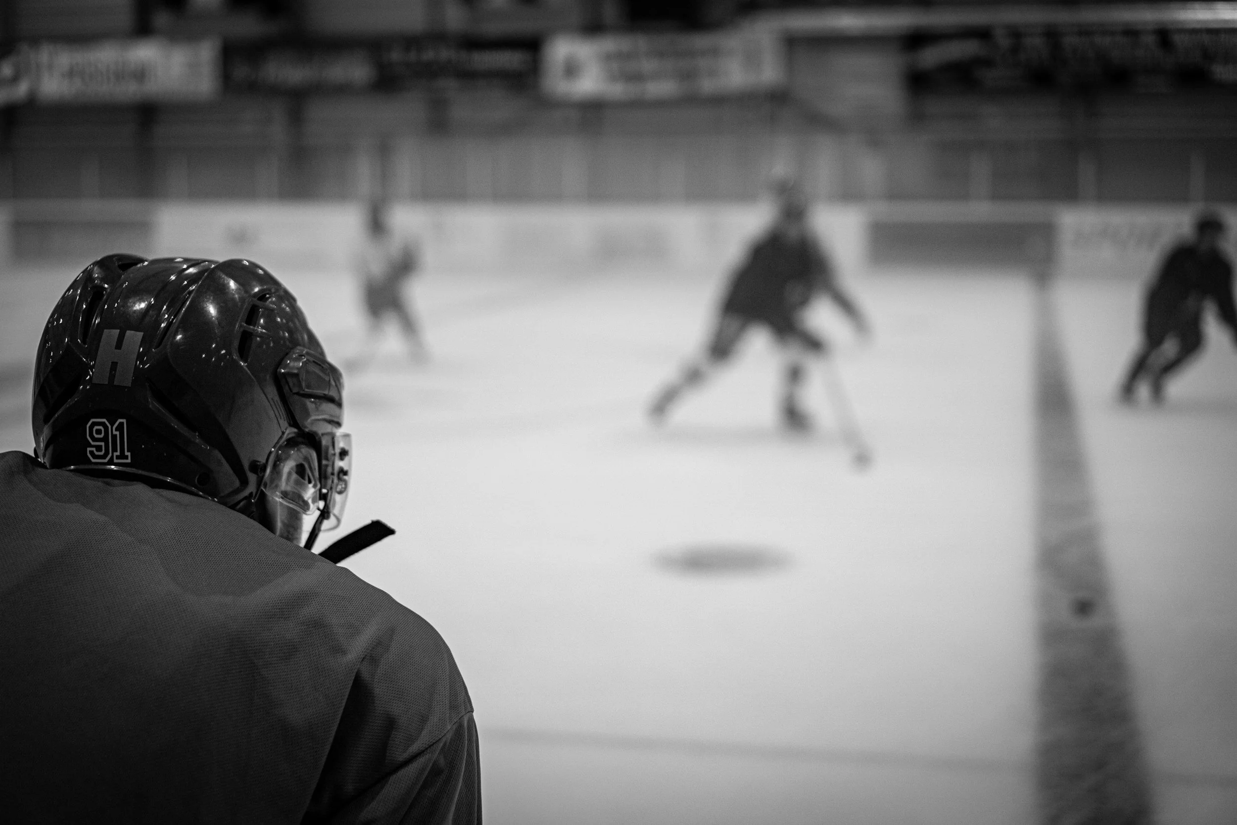 A hockey player sitting in the ice rink, looking towards the ice where other players are skating in the background.