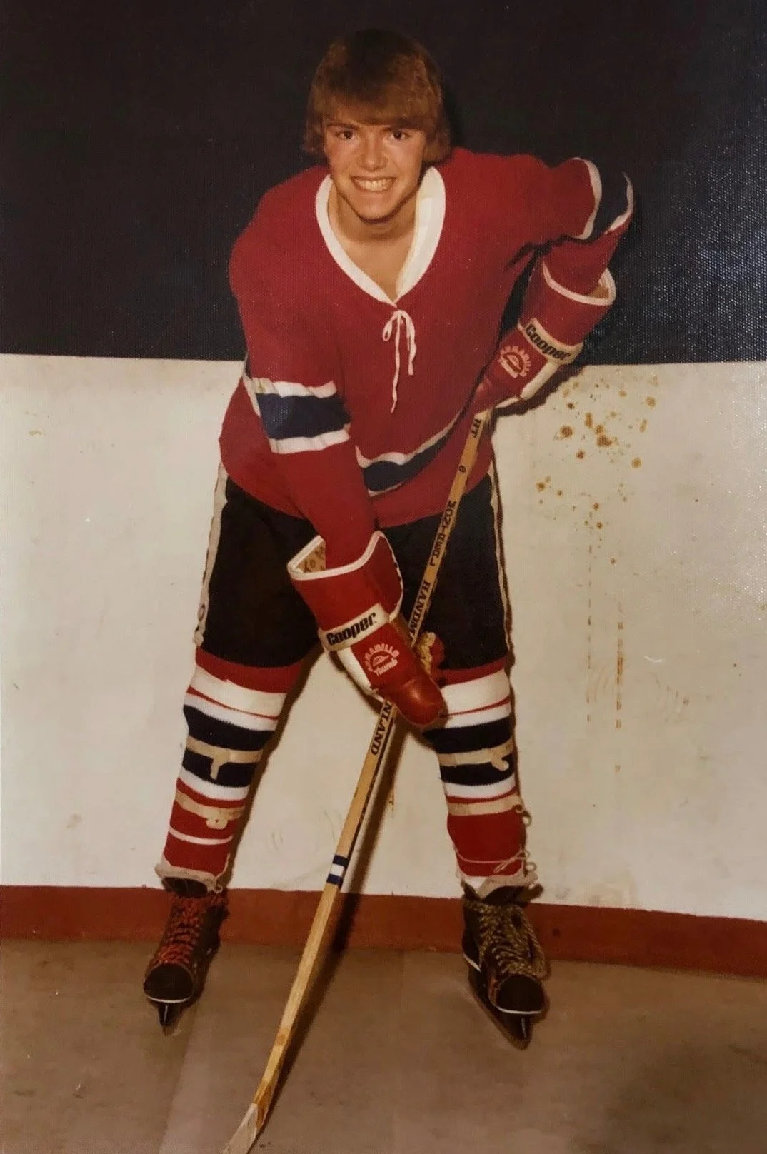 Young man in a red hockey jersey and skates, holding a hockey stick, standing on the ice with a black and white wall background.