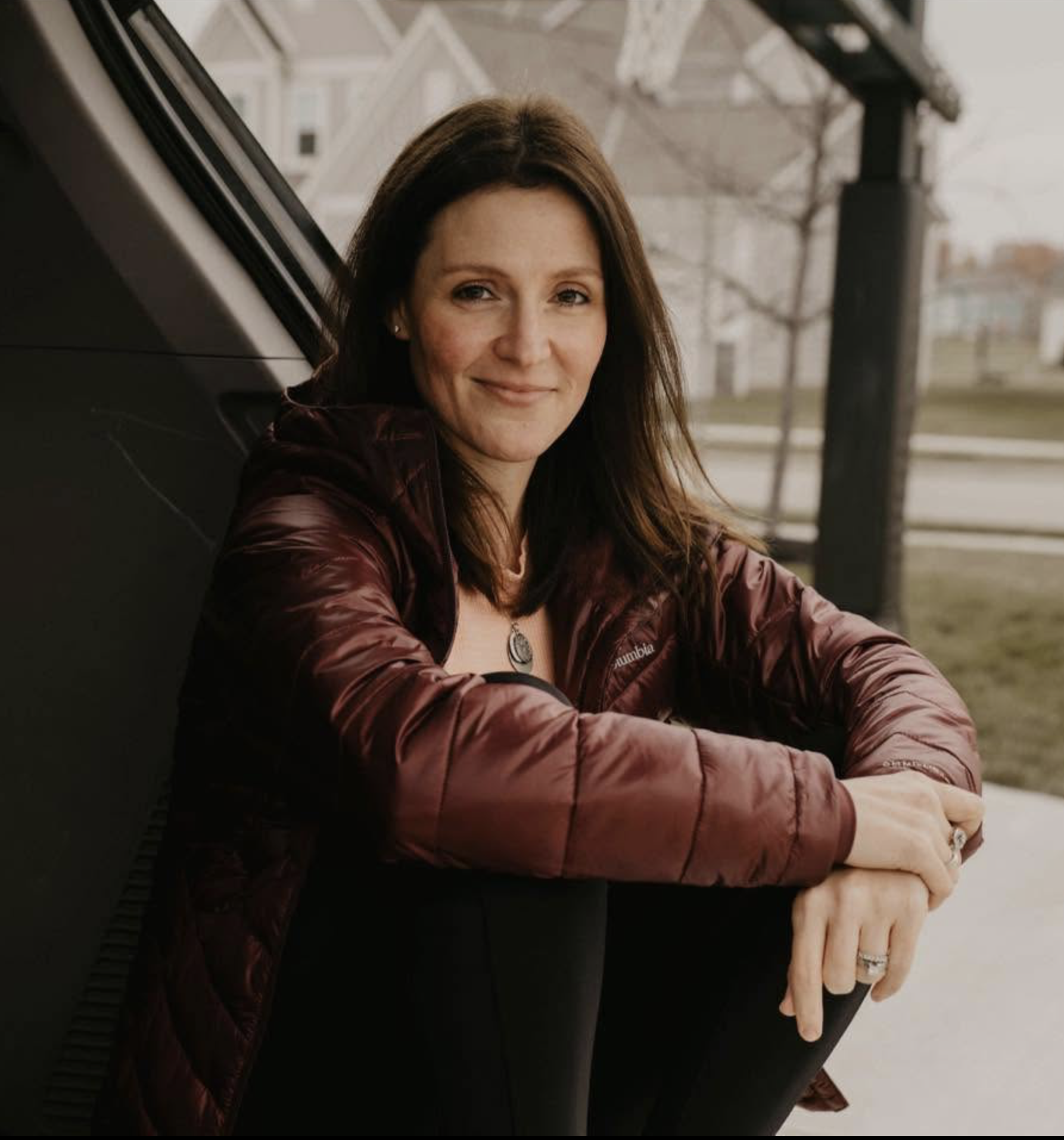 Close-up of a woman with long brown hair, smiling, wearing a maroon puffy jacket and a necklace, sitting outdoors near a city structure with a wire fence and buildings in the background.