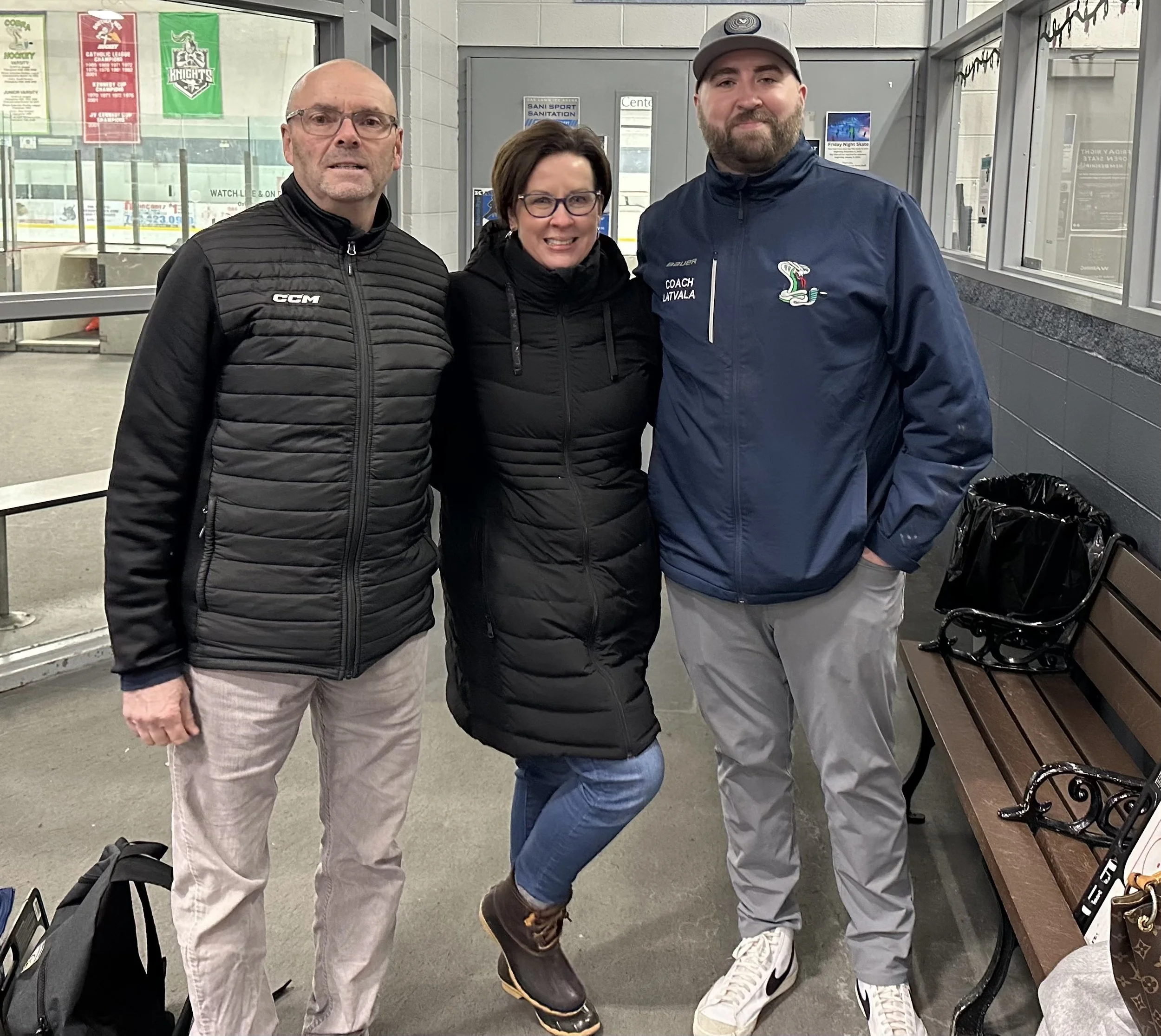 Three people standing together in an indoor ice skating rink, with a glass barrier and posters in the background, and bags on the floor beside them.