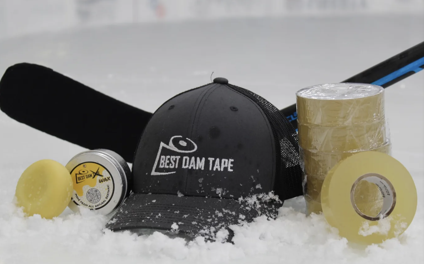Hockey equipment and supplies on ice, including a black baseball cap with white text reading 'Best Dam Tape,' yellow wax container, yellow wax ball, packing tape, and a hockey stick.