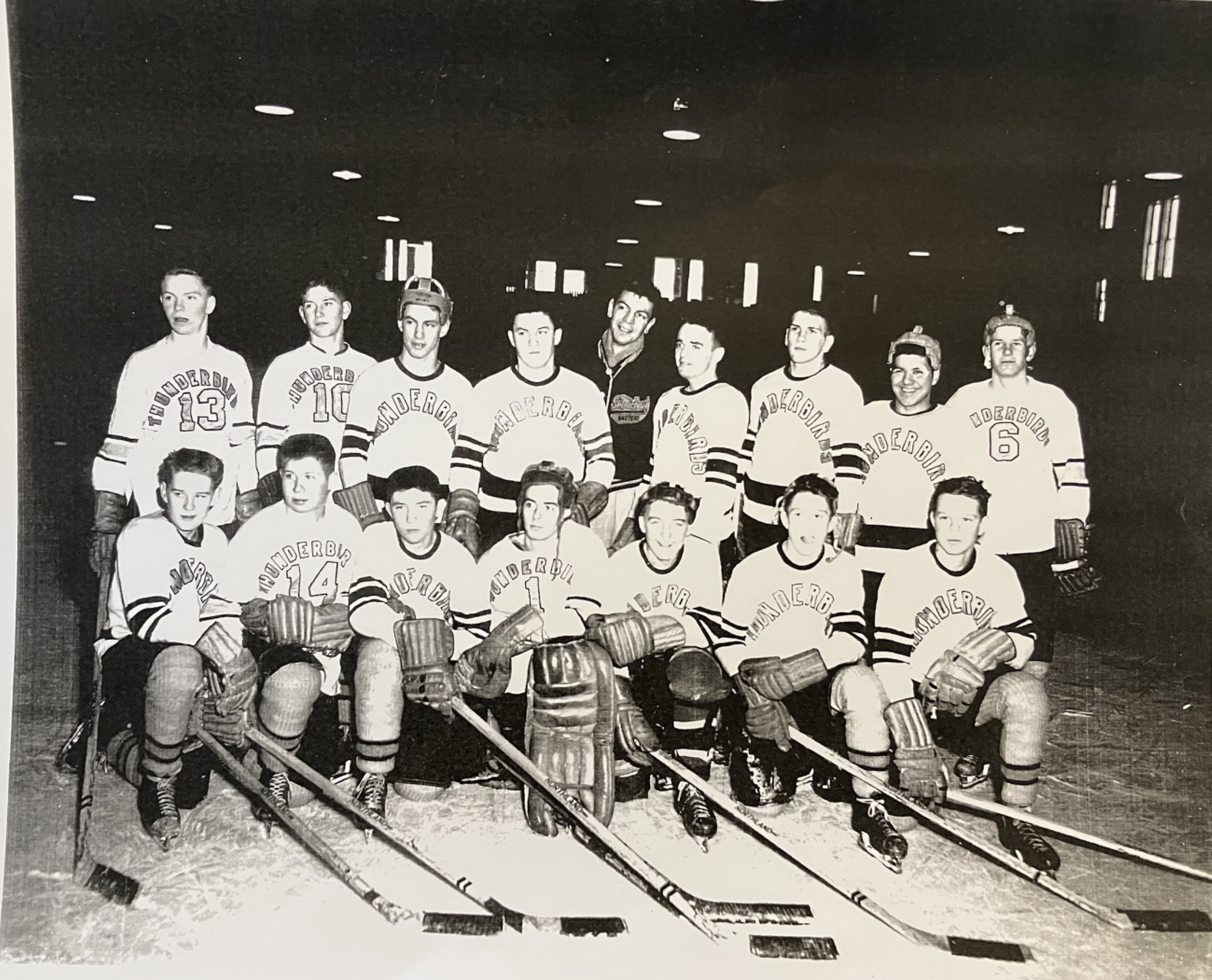 A black and white photo of a group of young male hockey players in uniform, holding hockey sticks, and standing in a line. Some are smiling, and a few are wearing helmets.