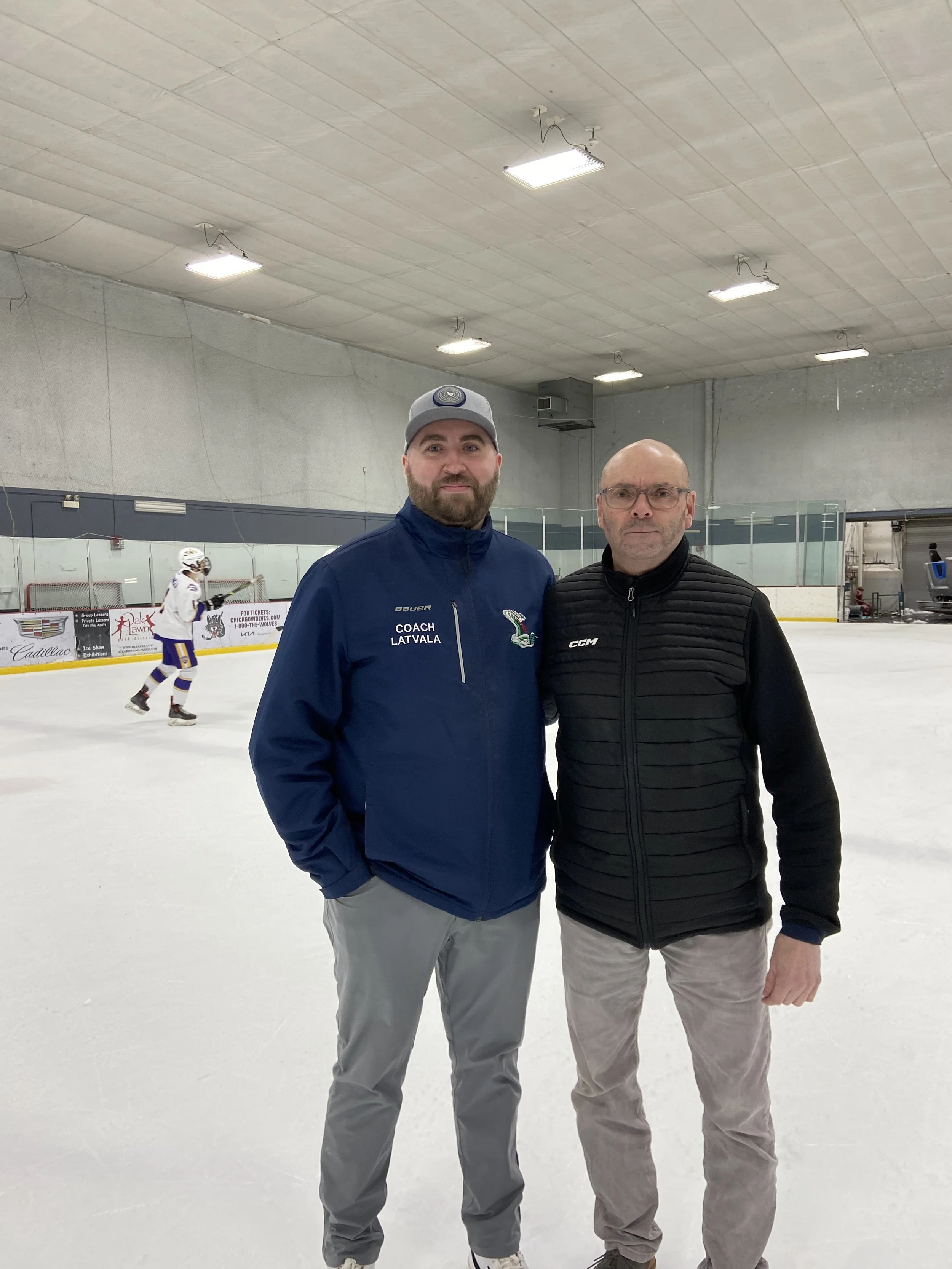 Two men standing on an ice rink with a hockey player skating in the background. They are wearing casual jackets and pants and are looking at the camera.
