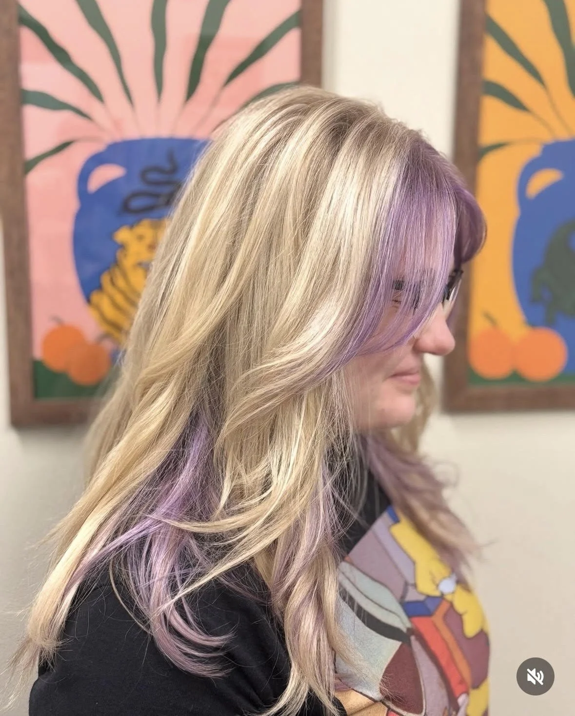 Side profile of a woman with blonde hair with purple highlights, wearing glasses, standing in front of colorful abstract paintings.