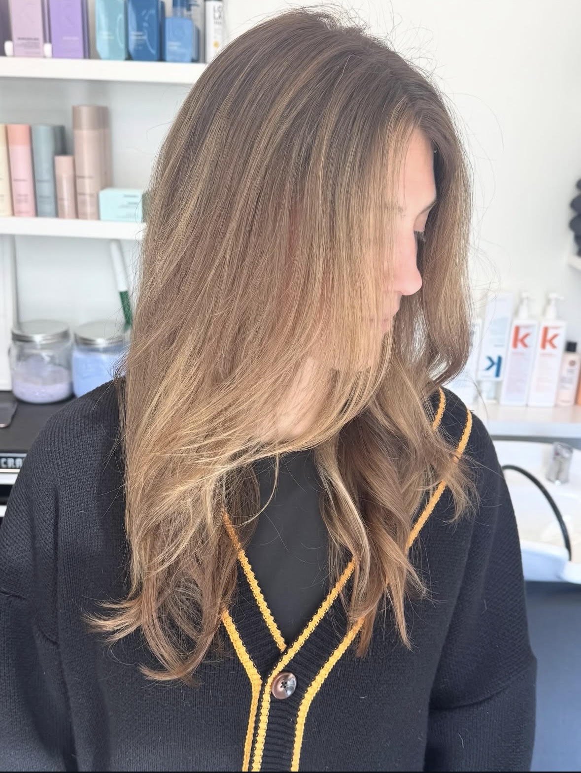 A woman with long, wavy, honey-colored hair standing in a salon with hair care products on shelves behind her.