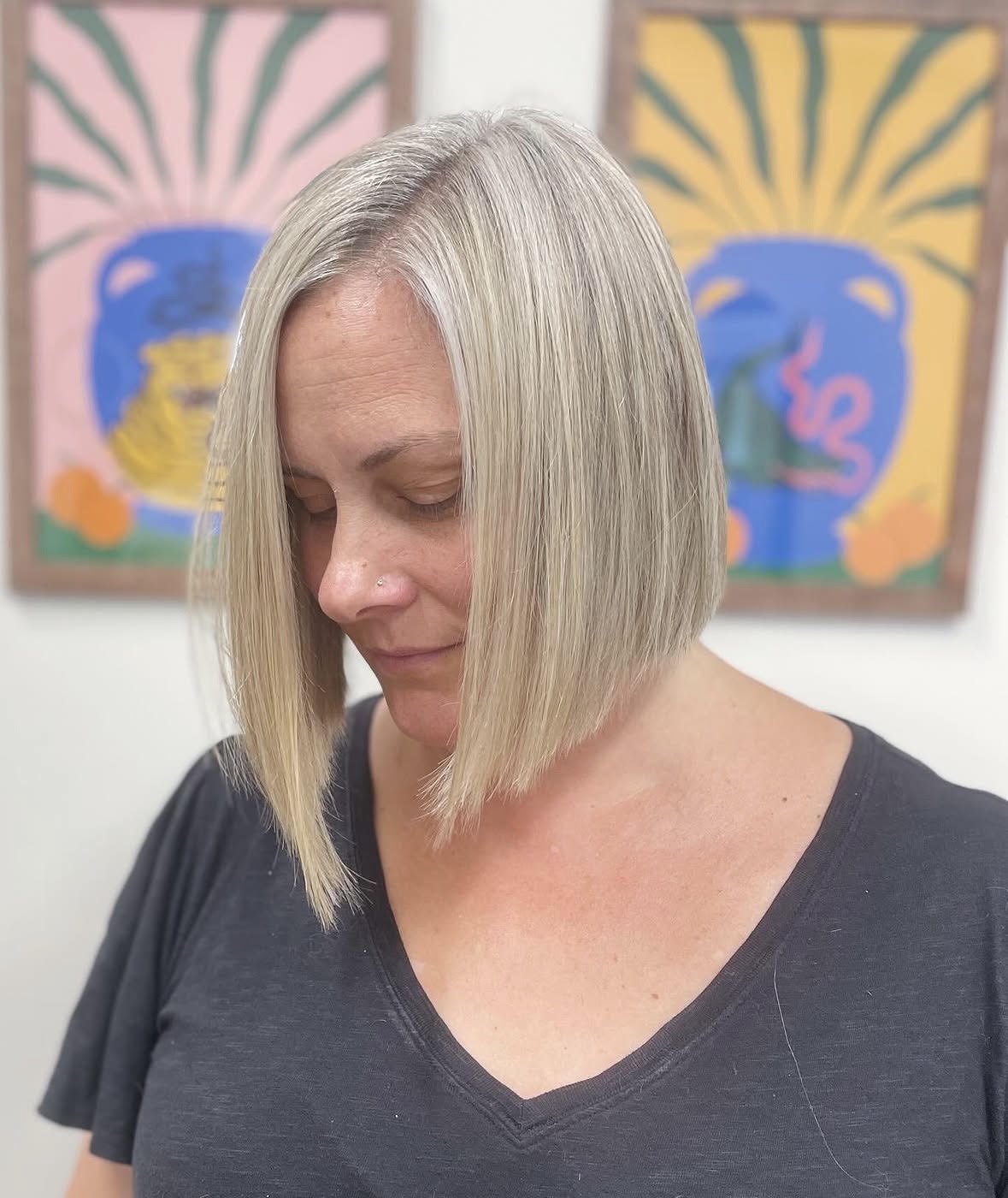 A woman with straight, blonde bob haircut looking down with eyes closed, standing in front of colorful abstract paintings.