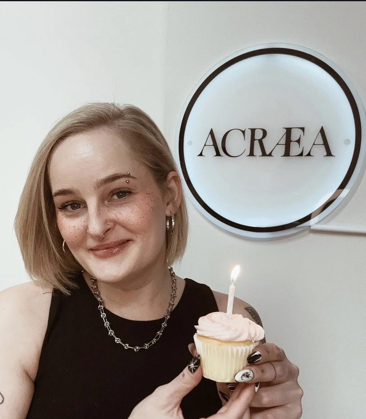 A woman with short blonde hair, tattoos, and multiple facial piercings, smiling while holding a cupcake with lit birthday candle in front of a sign that says 'ACREA'.