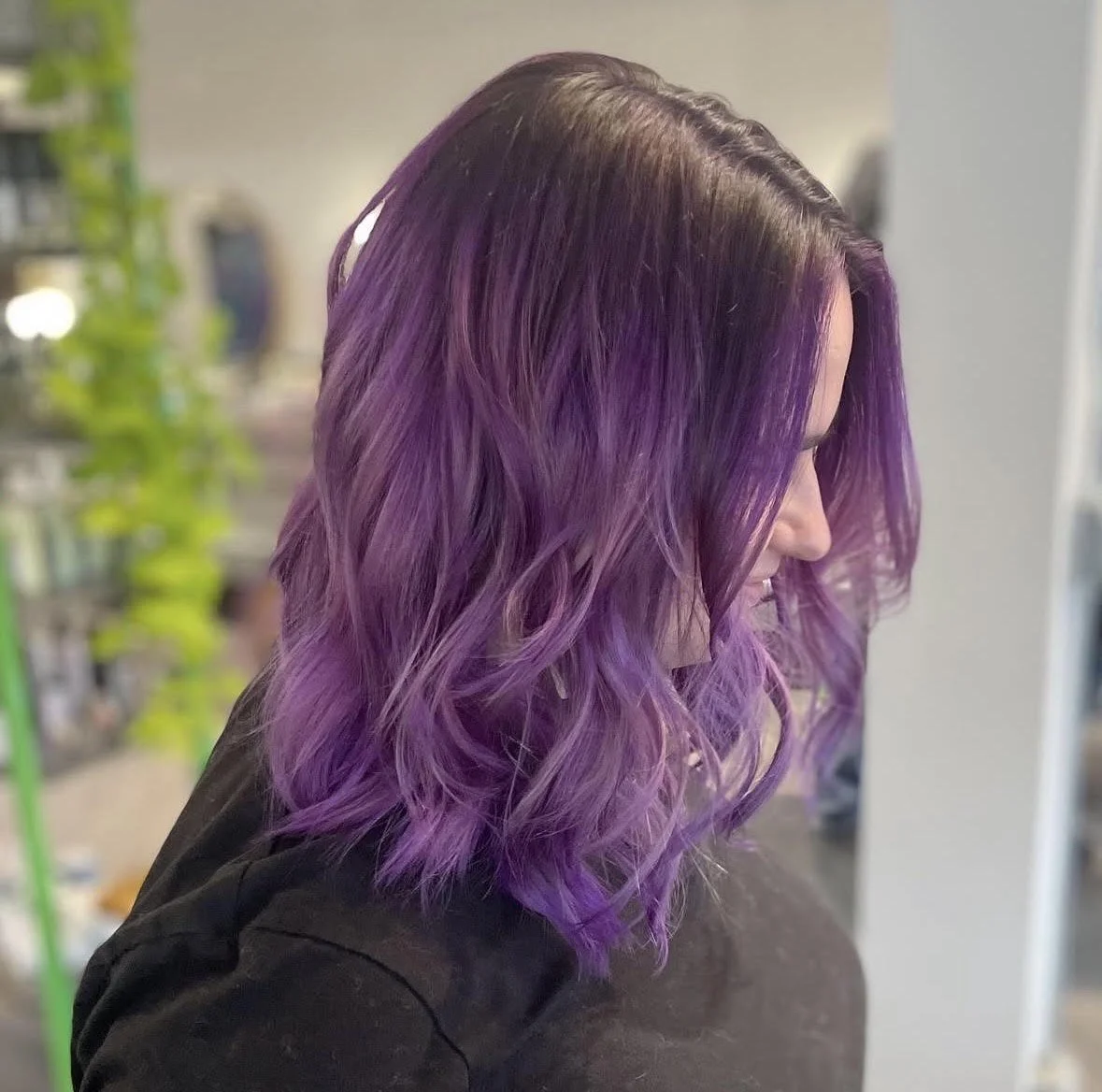 Side view of a woman with purple ombre wavy hair, smiling and wearing a black top.